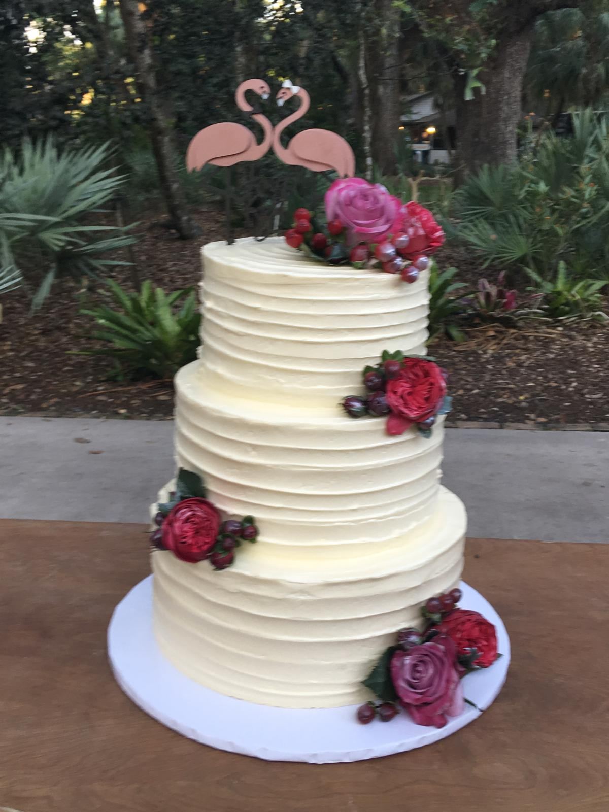 Three-tier buttercream cake with roses and flamingo topper in a garden