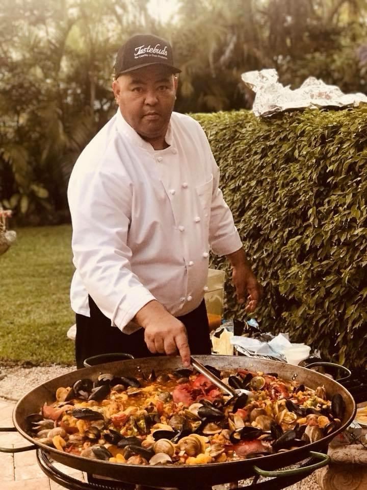 Chef in a Tastebuds cap serving seafood paella from a large pan outdoors